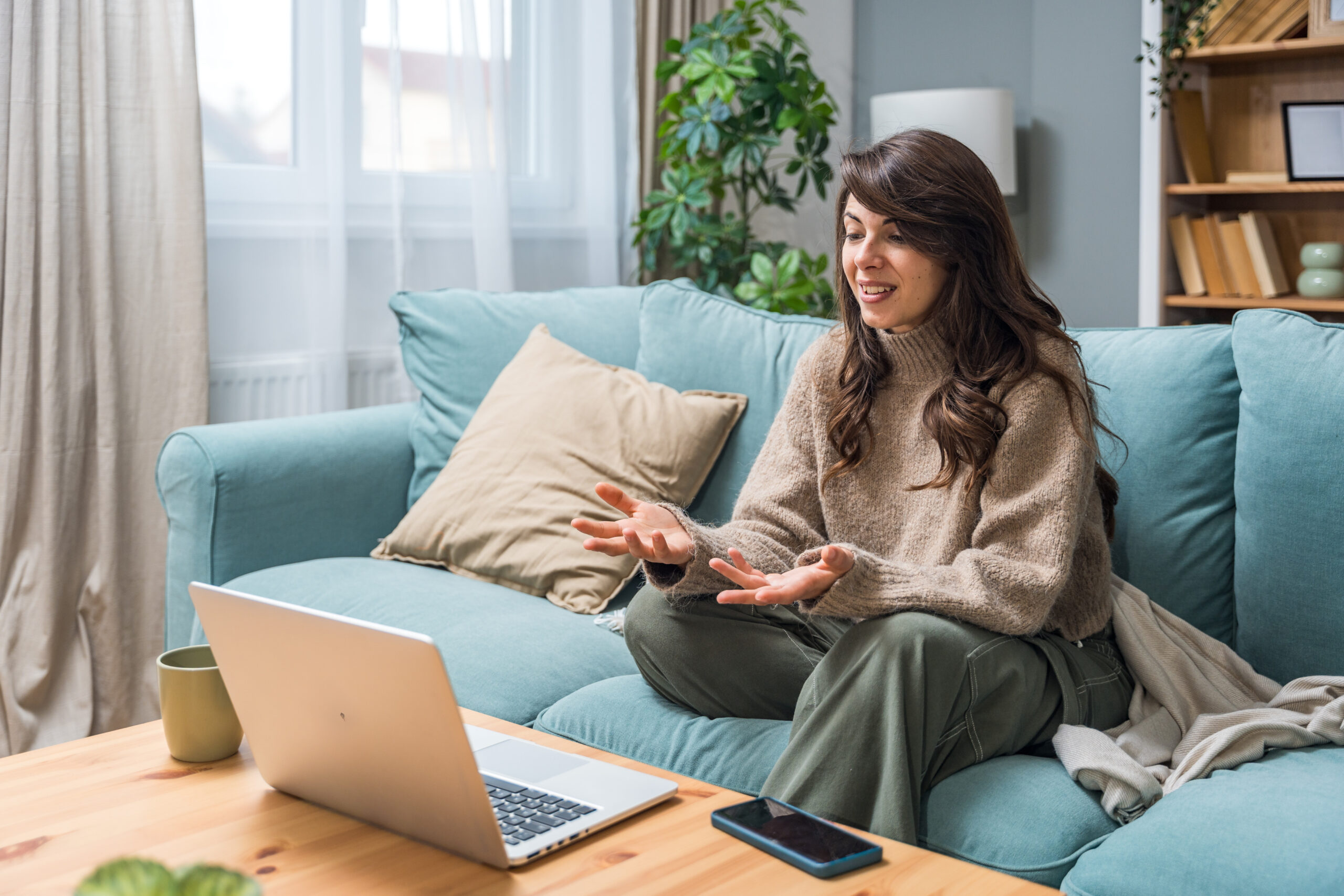 Young woman sitting on sofa during an online video call, smiling and touching her chest while speaking. Expressing emotion and connection through virtual communication.